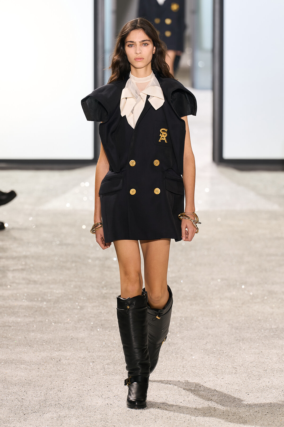 Model walking the runway in a black double-breasted mini dress with gold buttons, oversized shoulders, and a beige bow tie at the neck, paired with tall black boots and bracelets.
