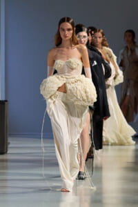 Model walks the runway in a cream corset with floral bust and a voluminous textured skirt, gloves, and long satin pants texture visible on the floor-styled look.