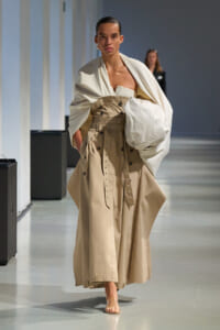 Model walking a runway in a tan trench dress with a wide belt and a large white draped cape.