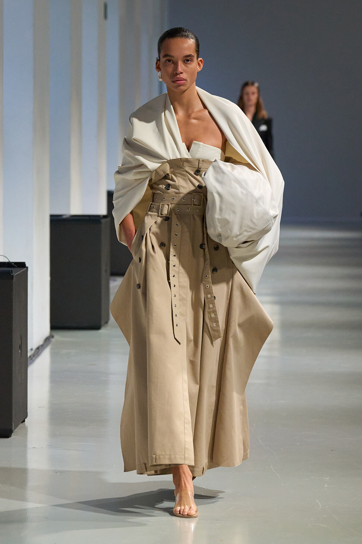 Model walking a runway in a tan trench dress with a wide belt and a large white draped cape.