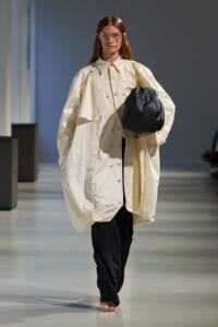 Model on a runway wearing an oversized cream trench coat, black pants, and carrying a large black bag.
