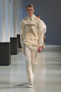 Model walking down a runway in an all-cream ensemble with layered ruffled peplum and structured jacket.