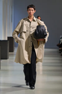 Model walking a runway in an oversized beige trench coat, black pants, carrying a large black leather bag under the arm.
