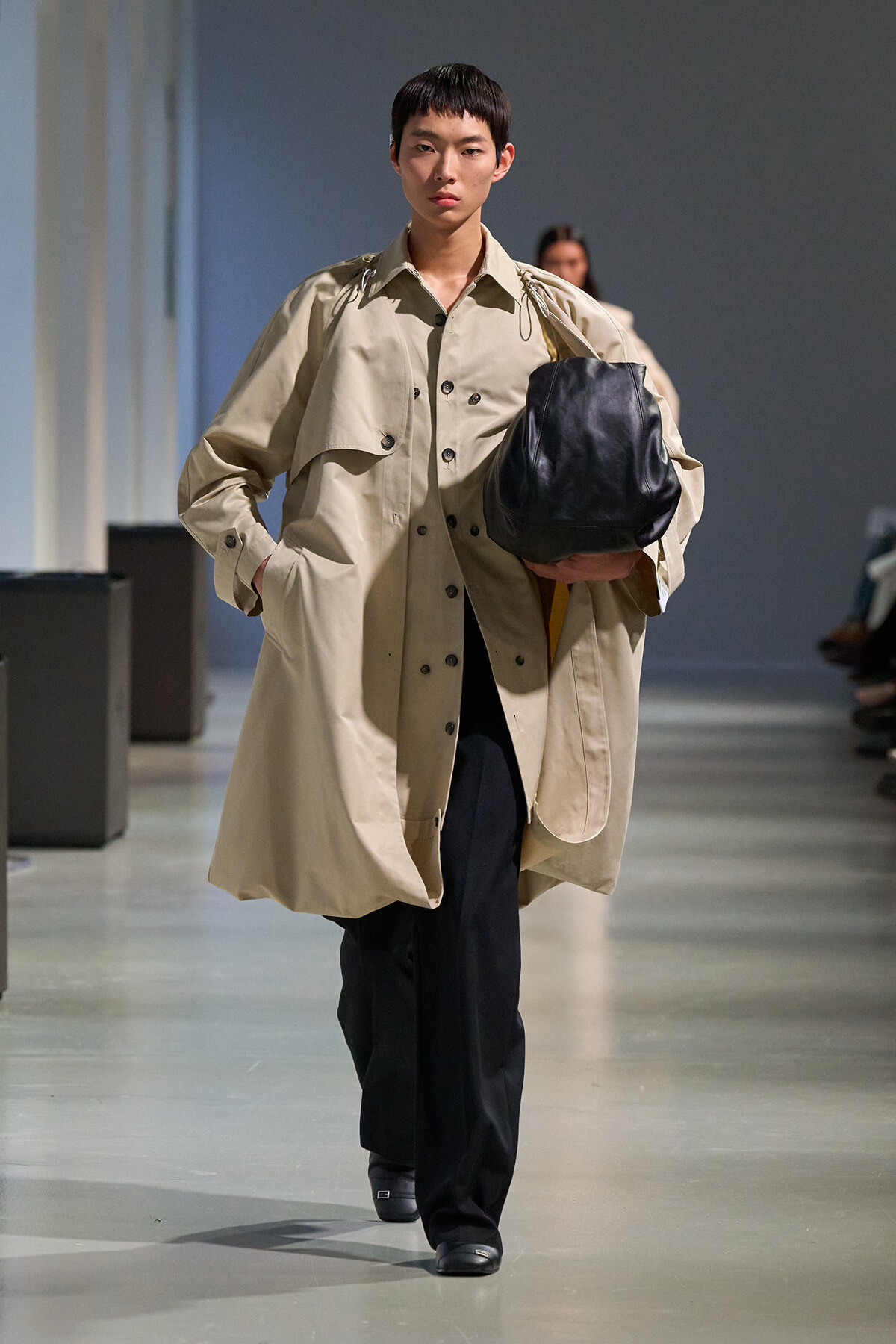 Model walking a runway in an oversized beige trench coat, black pants, carrying a large black leather bag under the arm.