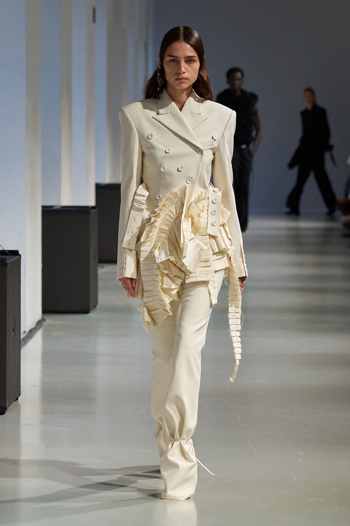 Model on a runway wearing a cream double-breasted suit with dramatic ruffled fabric at the waist.