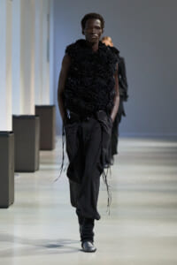 Male model walking a runway in an all-black outfit with a textured, flower-like top and loose black pants, hands in pockets.