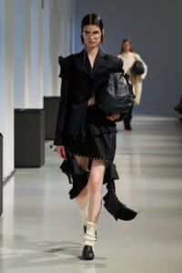 Model in a black ruffled ensemble walks a runway, carrying a large black bag and wearing statement sunglasses with dangling chains.