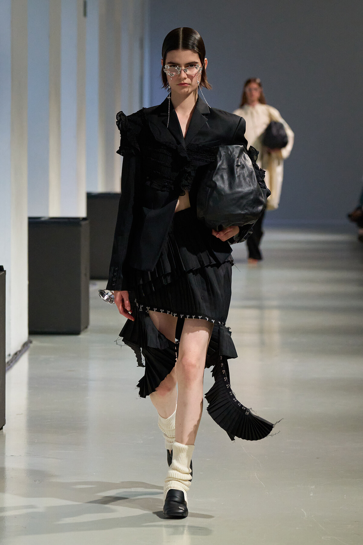 Model in a black ruffled ensemble walks a runway, carrying a large black bag and wearing statement sunglasses with dangling chains.