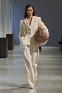 Model walking a runway in an oversized cream suit, holding a brown slouchy handbag.