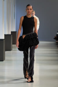 Model on a runway wearing a fitted black sleeveless top and a voluminous, ruffled black skirt over dark pants with silver embroidery. She wears black sandals and a bold earring, walking toward the camera.