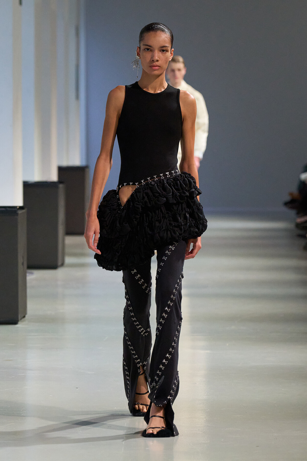 Model on a runway wearing a fitted black sleeveless top and a voluminous, ruffled black skirt over dark pants with silver embroidery. She wears black sandals and a bold earring, walking toward the camera.