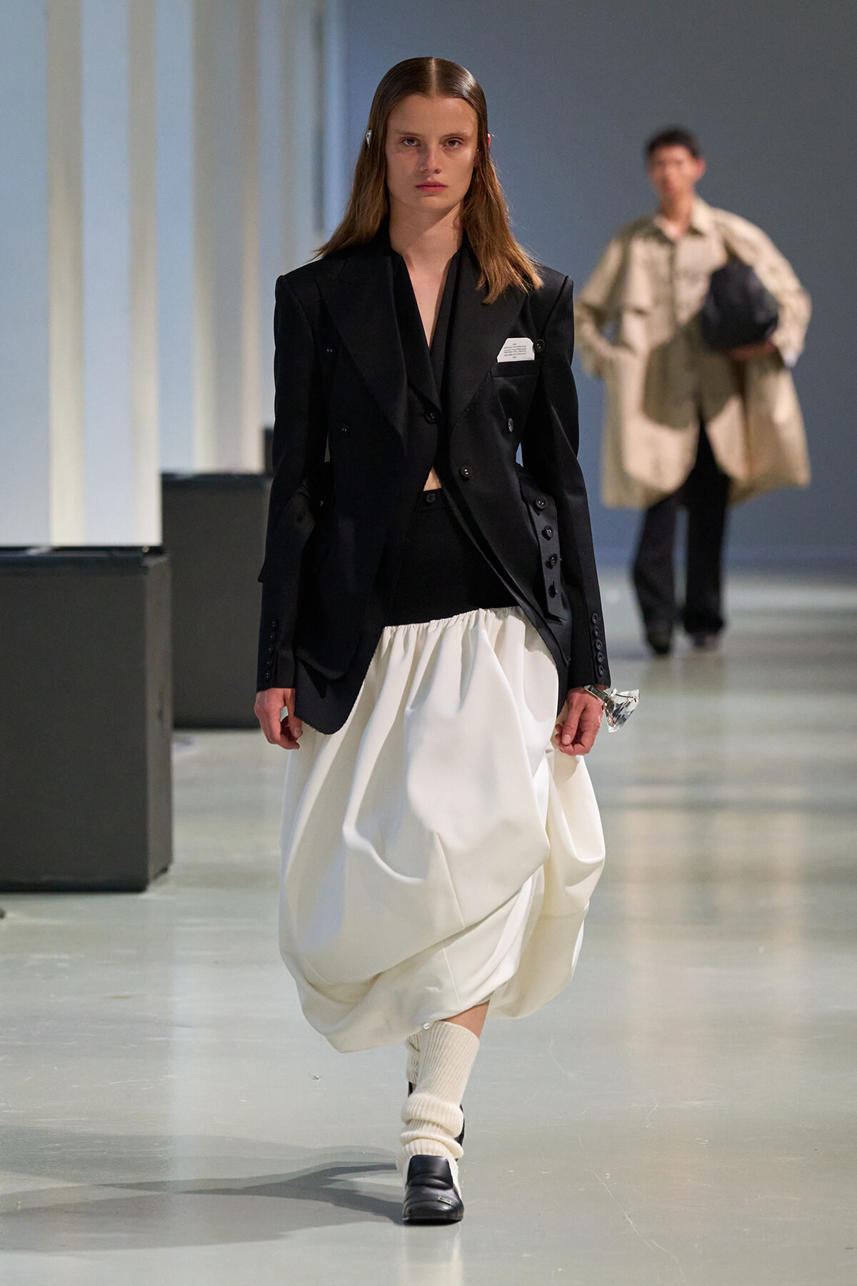 Model on a runway wearing a black structured blazer paired with a voluminous white draped skirt.