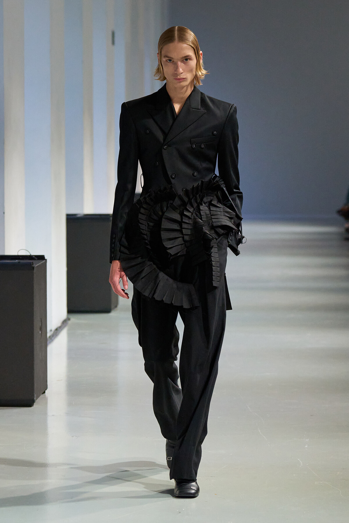 Model walking the runway in a black double-breasted suit with a large, sculpted pleated ruffle at the waist.