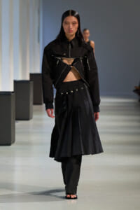 Runway model in a black avant-garde outfit: cropped jacket with zipper details and a high-waisted pleated skirt showing midriff.
