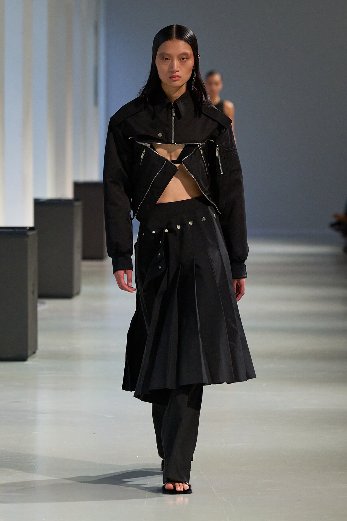 Runway model in a black avant-garde outfit: cropped jacket with zipper details and a high-waisted pleated skirt showing midriff.
