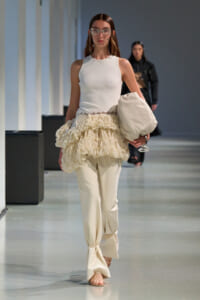 Runway model in a white sleeveless top with a cream ruffled peplum over slim pants, carrying a large beige bag and wearing crystal sunglasses.