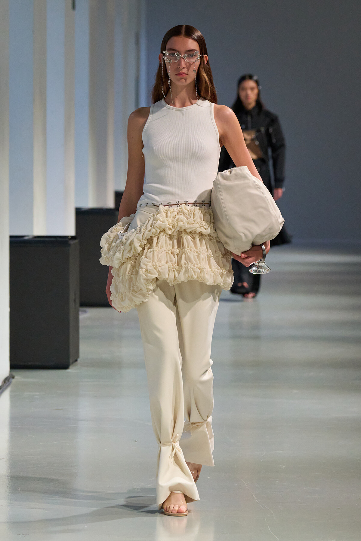 Runway model in a white sleeveless top with a cream ruffled peplum over slim pants, carrying a large beige bag and wearing crystal sunglasses.