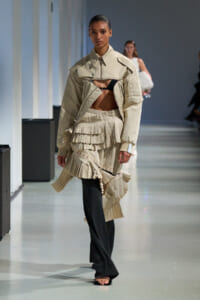 Runway model in a beige cropped jacket with exposed midriff, paired with a layered pleated beige skirt and black pants underneath.