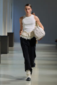 Male runway model walking the catwalk in a white sleeveless top, cream pleated belt, black pants, cream socks, black shoes, carrying a white bag.