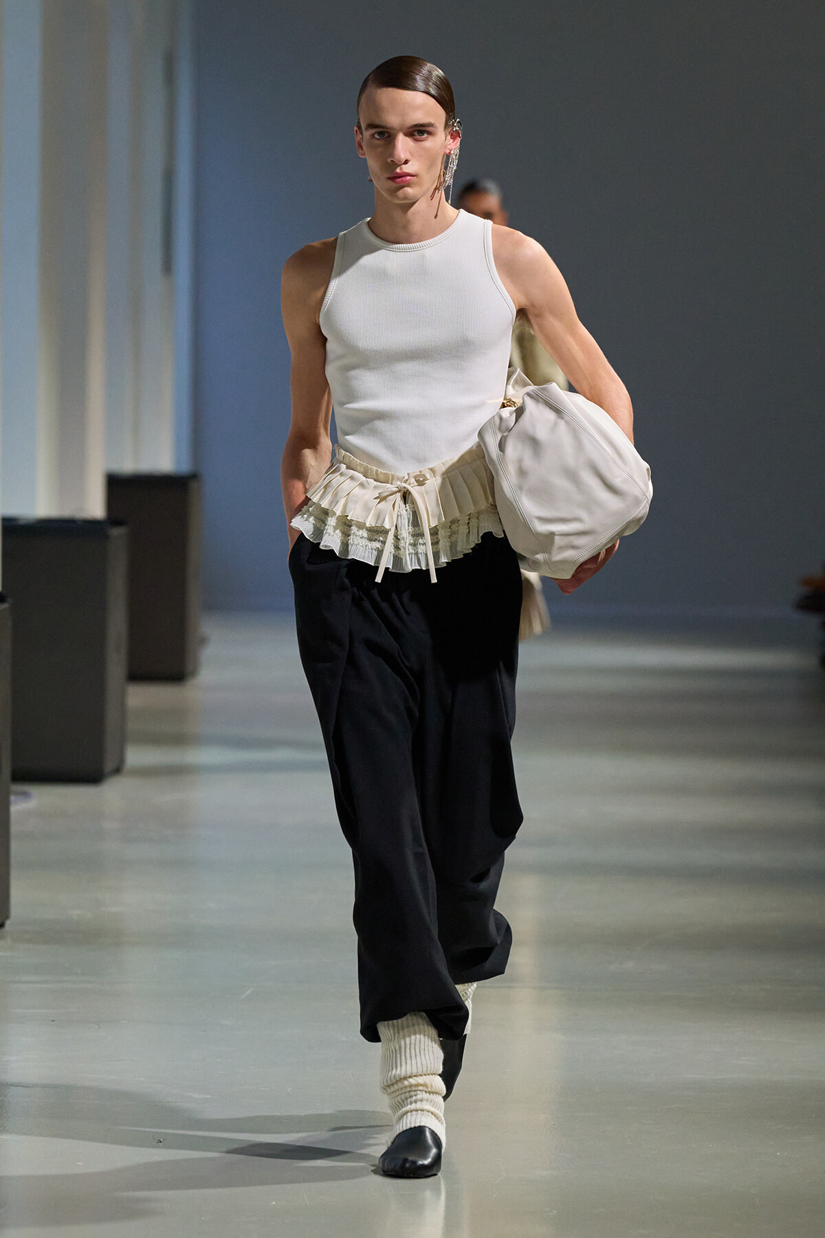 Male runway model walking the catwalk in a white sleeveless top, cream pleated belt, black pants, cream socks, black shoes, carrying a white bag.