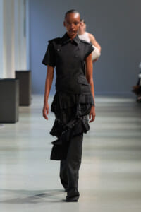 Model walking down a runway in a black, structured top with double-breasted buttons and layered, ruffled skirt accessories.