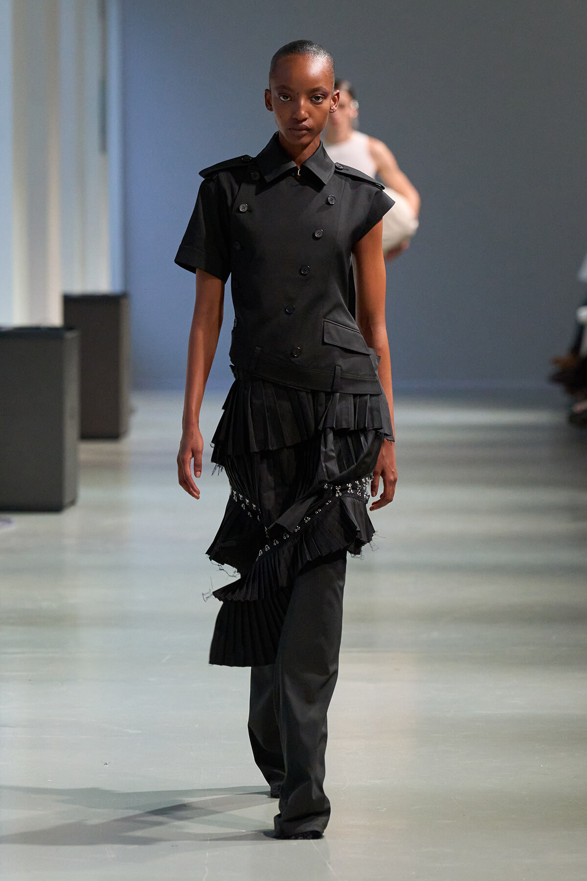 Model walking down a runway in a black, structured top with double-breasted buttons and layered, ruffled skirt accessories.