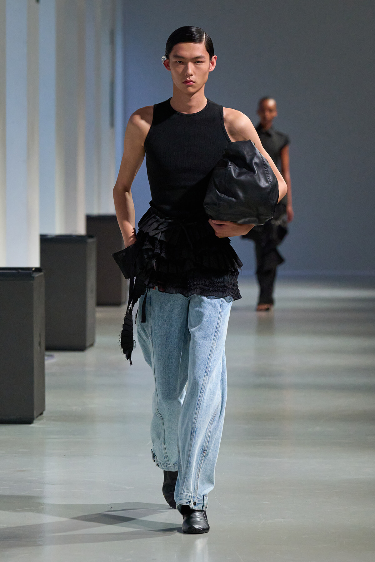 Model walks a runway in a black sleeveless top, light denim jeans, and a large black bag slung under the arm.