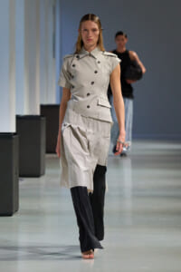 Female model walks the runway in a light gray, double-breasted trench top with an attached peplum over black pants, holding a crystal water bottle.
