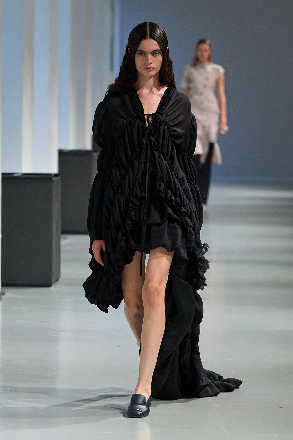 Model walking a fashion runway in a voluminous black ruffled dress with a long train and black loafers.