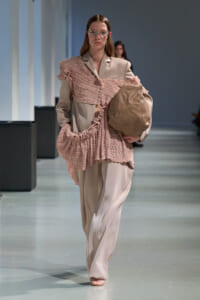 Model walking a runway in a beige suit with a pink, ruffled overlay and carrying a large taupe bag.