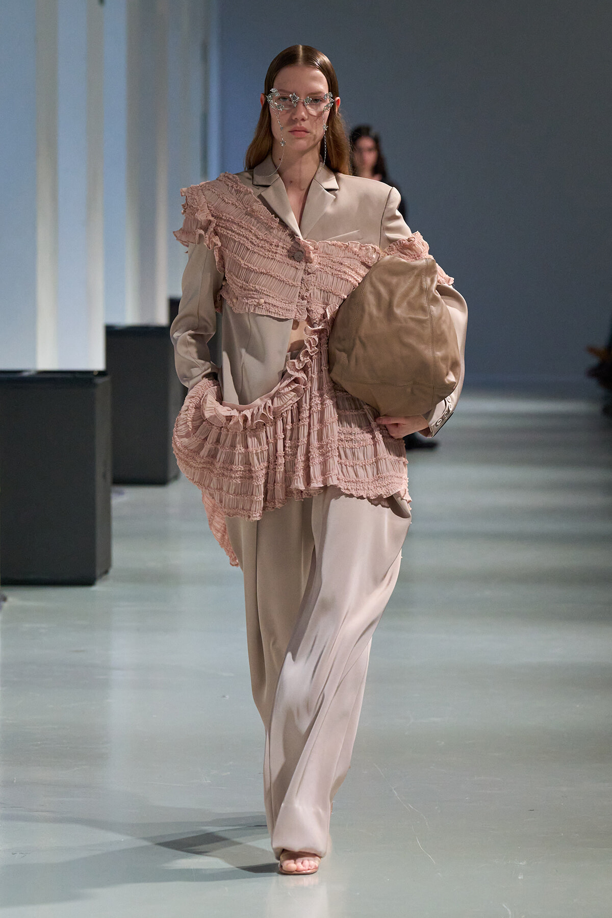 Model walking a runway in a beige suit with a pink, ruffled overlay and carrying a large taupe bag.