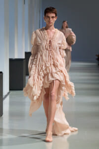 Model walking a runway in a pale pink, ruffled, voluminous dress with a gathered waist and tie-front details.