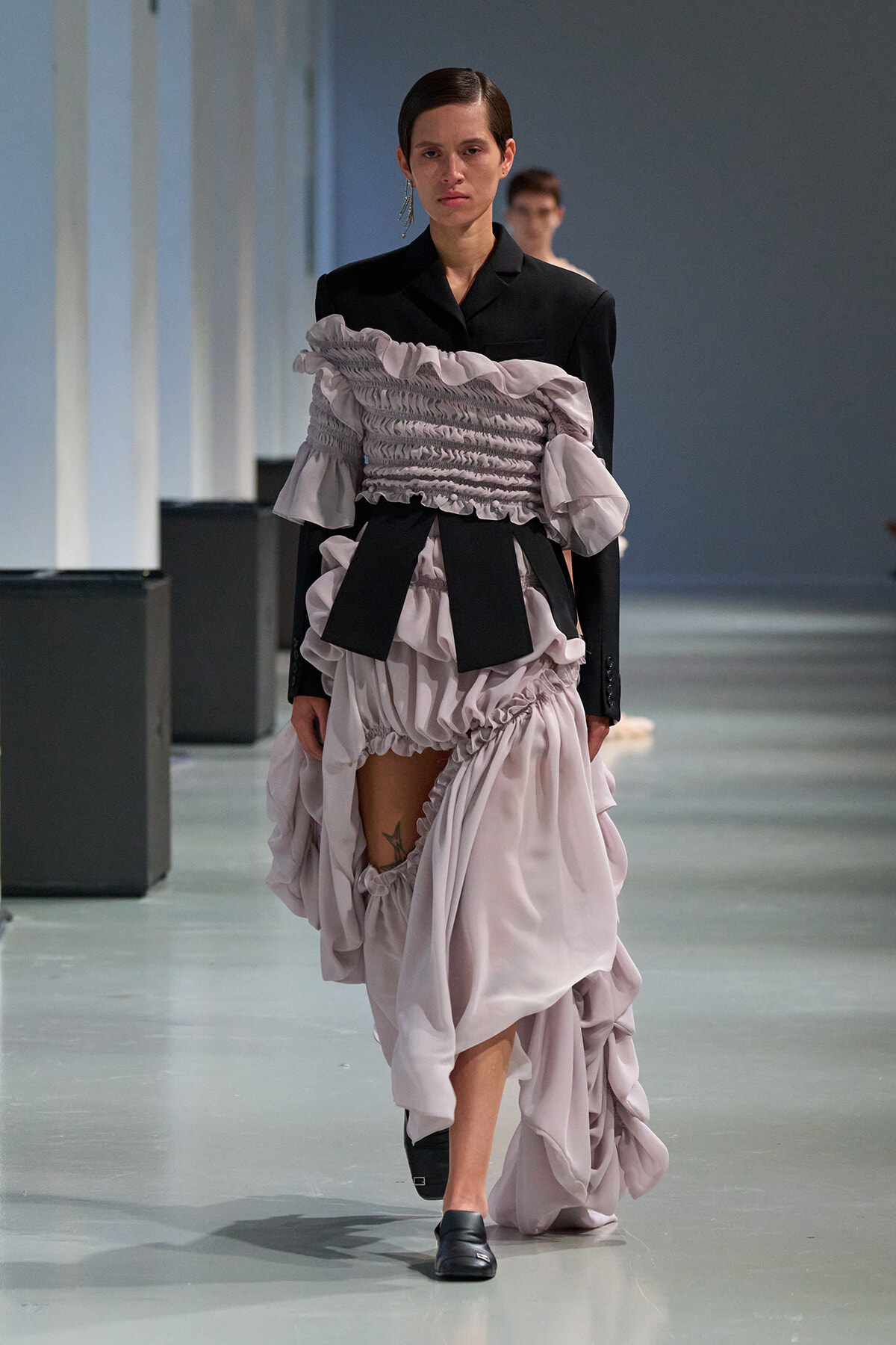 Model walking a runway in a black blazer paired with a lavender ruched, ruffled gown that reveals a leg tattoo and black loafers.