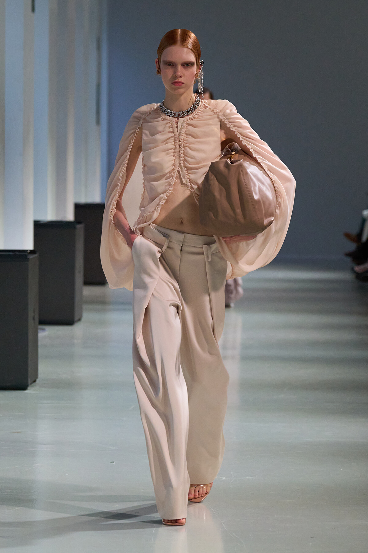 Model on a runway wearing a pale pink ruched blouse with cape-like sleeves and wide-leg beige pants, carrying a large taupe bag.