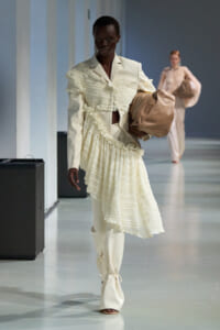 Model walks a fashion runway in an ivory suit with a tiered, ruffled overlay, holding a tan handbag.