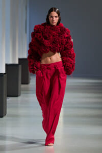 Female model walks a fashion show wearing a cropped, voluminous red ruffled sweater with matching red wide-leg pants, barefoot on the catwalk.