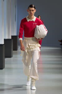 Male model on a runway wearing a red knit sweater with a white collar, cream pleated skirt over pants, and a large white bag in hand.