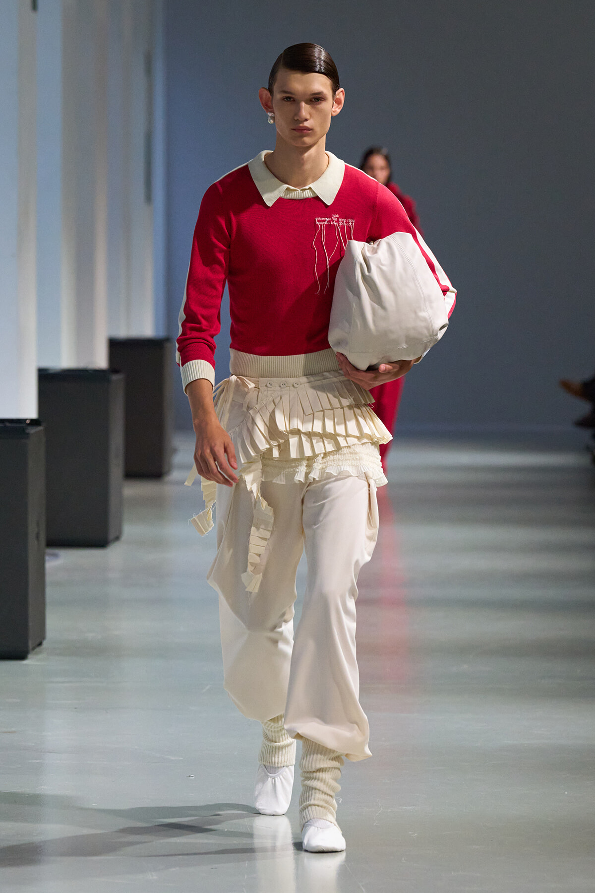 Male model on a runway wearing a red knit sweater with a white collar, cream pleated skirt over pants, and a large white bag in hand.