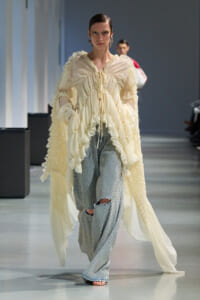 Model on a runway wearing a cream ruffled blouse with a long sheer cape and light-blue dotted pants with knee holes.
