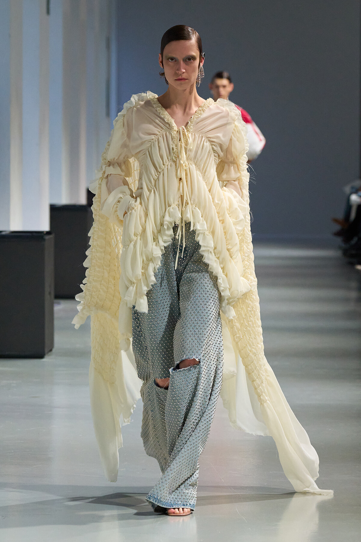 Model on a runway wearing a cream ruffled blouse with a long sheer cape and light-blue dotted pants with knee holes.