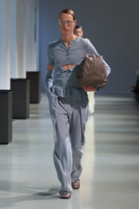 Model walking a runway in a pale blue cropped cardigan with cutouts, matching high-waisted pants, and a large brown tote bag.