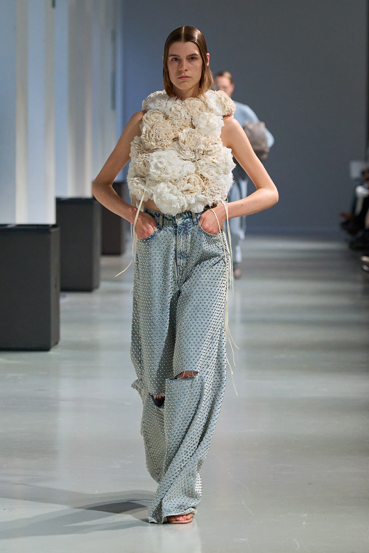 Model walks a runway wearing a voluminous, cream floral-constructed top with exposed denim jeans featuring knee holes; fashion-show setting.
