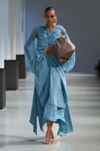 Model on a runway wearing a sky-blue, ruched gown with oversized, cape-like sleeves and a cutout bodice, carrying a taupe/gray clutch.