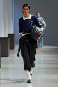 Model walking a runway in a navy sweater with white collar, black layered skirt, white leg warmers, and black shoes, carrying a gray bag