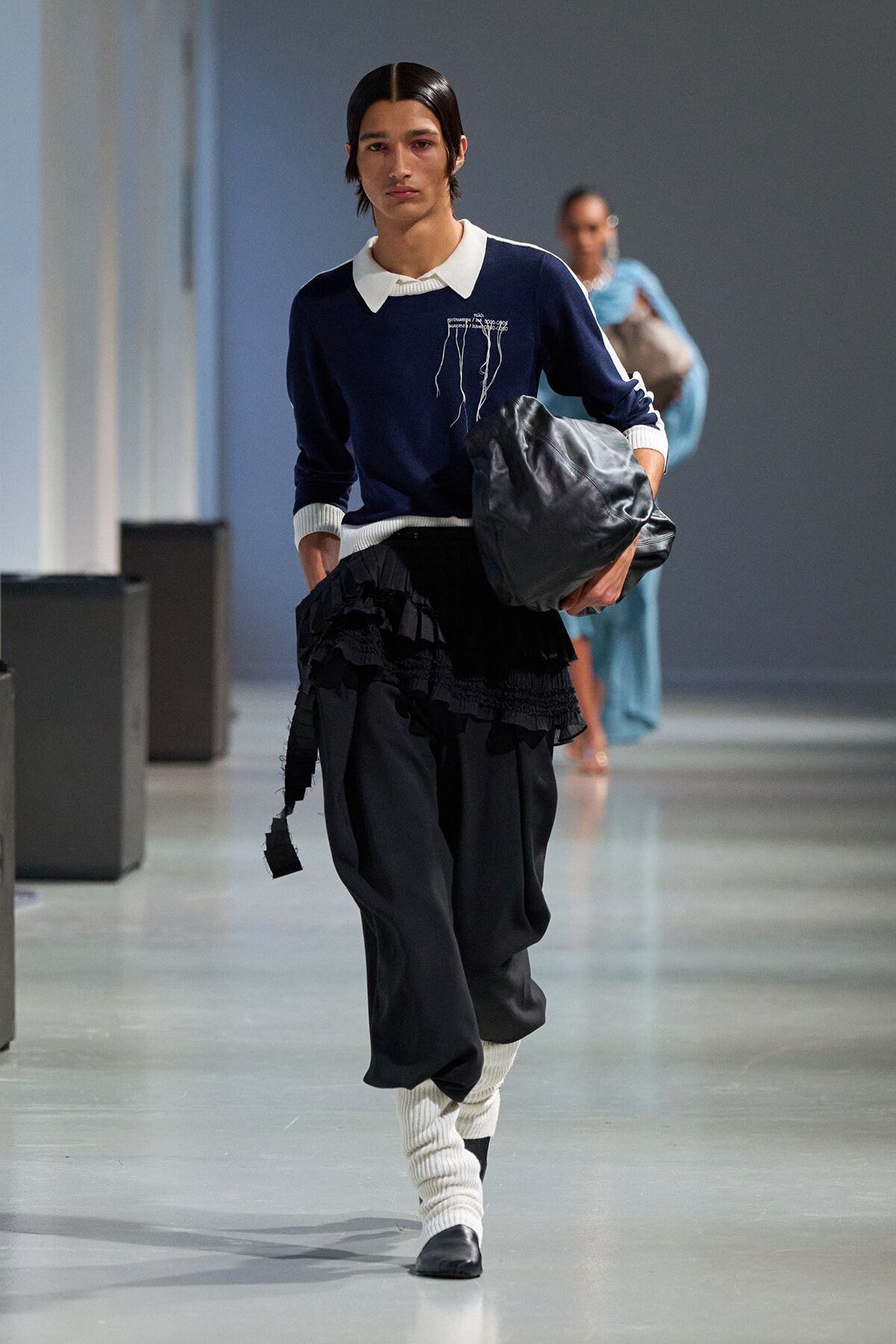 Model walking a runway in a navy sweater with white collar, black layered skirt, white leg warmers, and black shoes, carrying a gray bag