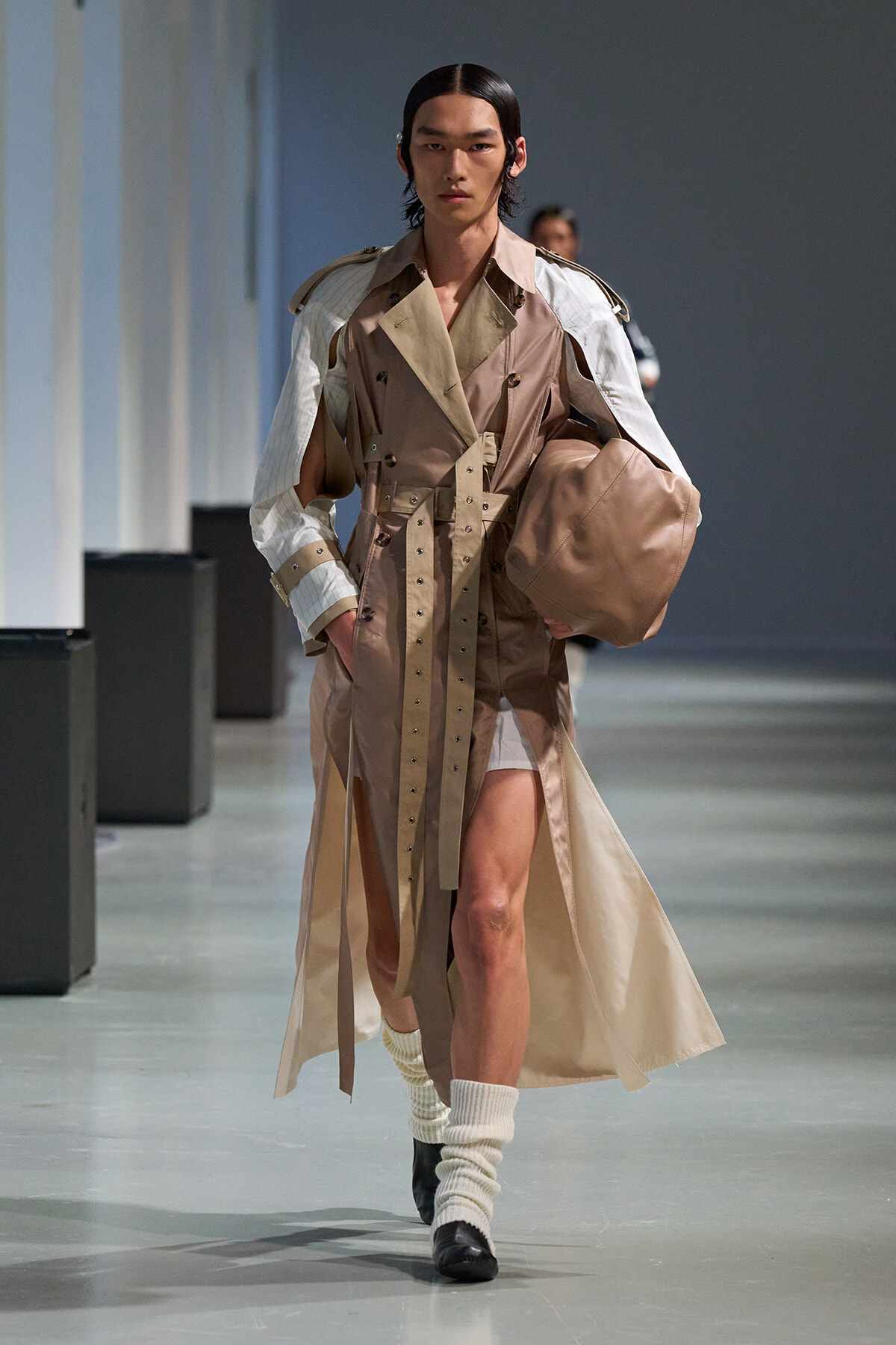 Model walking the runway in a tan-beige trench coat with a wide belt, oversized layered sleeves, white leg warmers, black shoes, and a brown bag