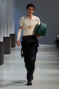 Male model walking a runway in a cream cropped cardigan with a cut-out midsection, black layered skirt, and black shoes, carrying a dark green bag.