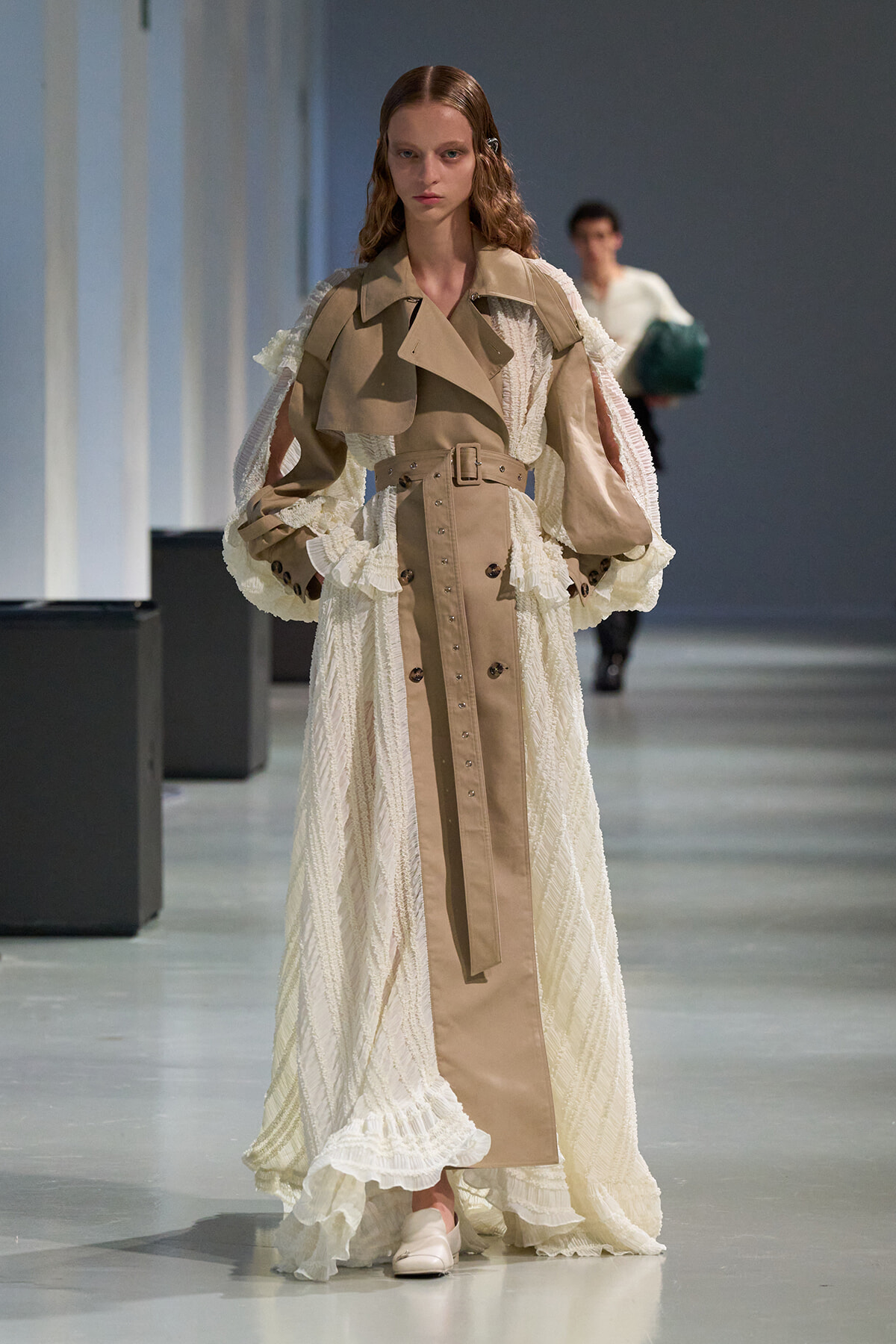 Model walks a runway wearing a beige belted trench layered over a white lace gown with voluminous sleeves and ruffles.