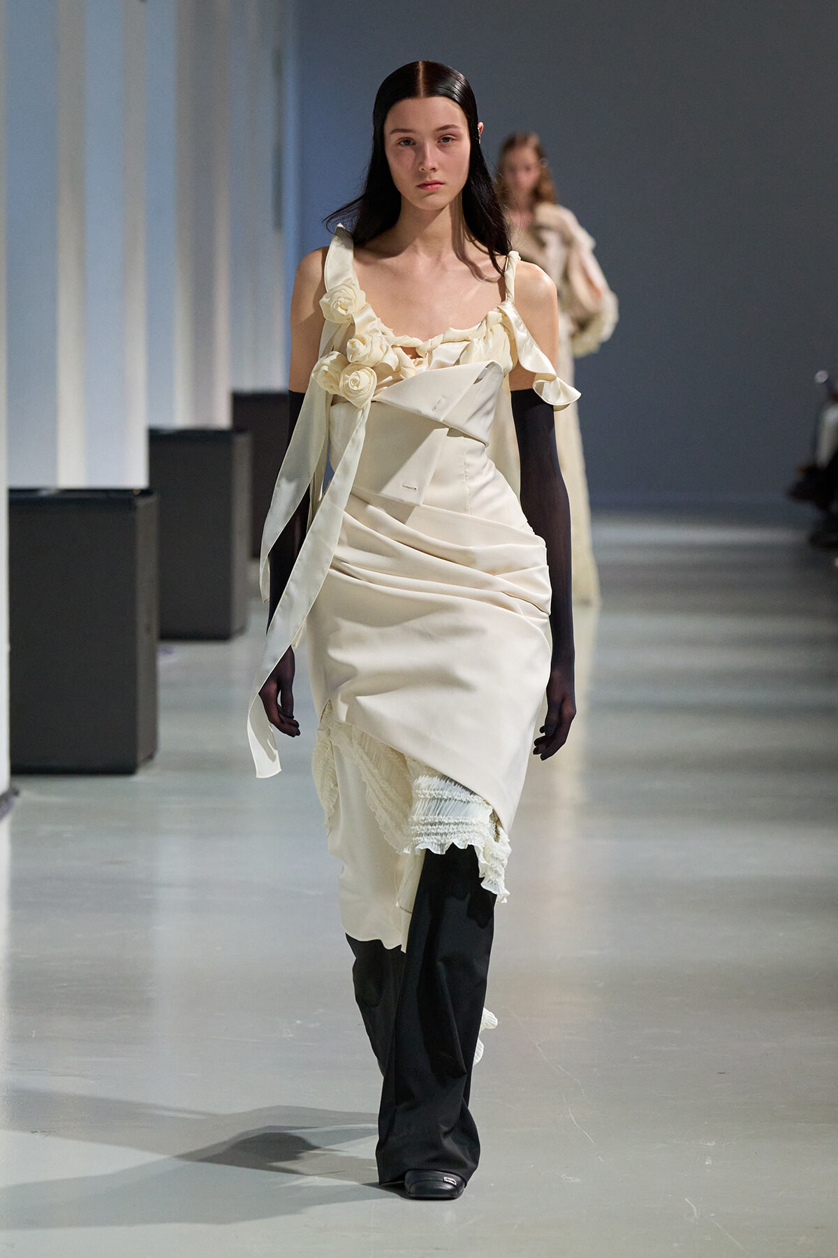 Model walking a runway in a cream draped dress with floral appliqués and long black gloves, neutral makeup, focused expression.