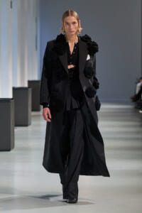 Model walks the runway in a long black coat adorned with oversized black floral appliqués, paired with black top and trousers for a monochrome look.